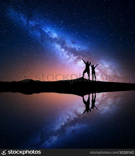 Milky Way. Night starry sky and silhouette of standing happy man and woman with raised up arms on the hill near the lake with sky reflection in water. Milky way with people on the mountain. Landscape