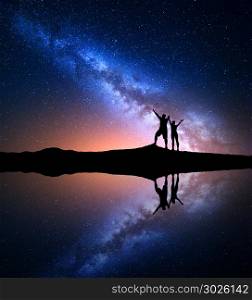 Milky Way. Night starry sky and silhouette of standing happy man and woman with raised up arms on the hill near the lake with sky reflection in water. Milky way with people on the mountain. Landscape