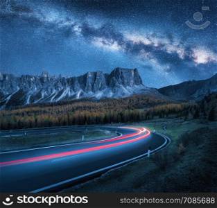Milky Way over mountain road at night. Blurred car headlights on winding road in autumn. Beautiful landscape with blue starry sky with milky way, light trails, rocks, trees and highway in fall. Space