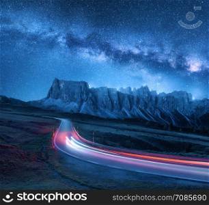 Milky Way over mountain road at night in summer. Blurred car headlights on winding road. Colorful landscape with sky with stars and blue milky way, light trails, rocks, trees and highway. Space