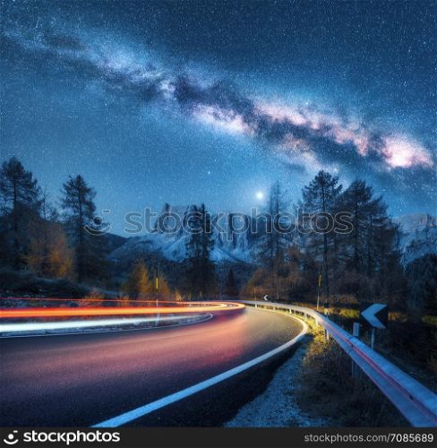Milky Way over mountain road. Blurred car headlights on winding road in autumn. Colorful night landscape with blue starry sky with milky way, moonlight, light trails, rocks, trees and highway. Space