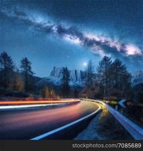 Milky Way over mountain road. Blurred car headlights on winding road in autumn. Colorful night landscape with blue starry sky with milky way, moonlight, light trails, rocks, trees and highway. Space