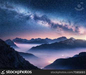 Milky Way over mountains in fog at night in summer. Landscape with foggy alpine mountain valley, purple low clouds, colorful starry sky with milky way, city illumination. Dolomites, Italy. Space