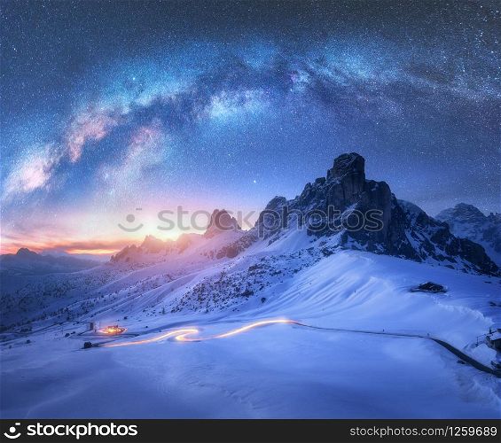 Milky Way over snowy mountains and blurred car headlights on the winding road at night in winter. Beautiful landscape with starry sky, snow covered rocks, house, roadway at sunset. Space and galaxy