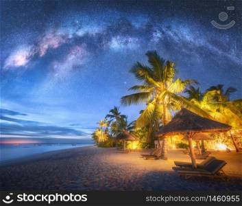 Milky Way over the sandy beach with green palm trees, sunbeds and umbrellas at night in summer. Landscape with sea coast, beautiful blue starry sky with galaxy. Travel in Zanzibar, Africa. Space