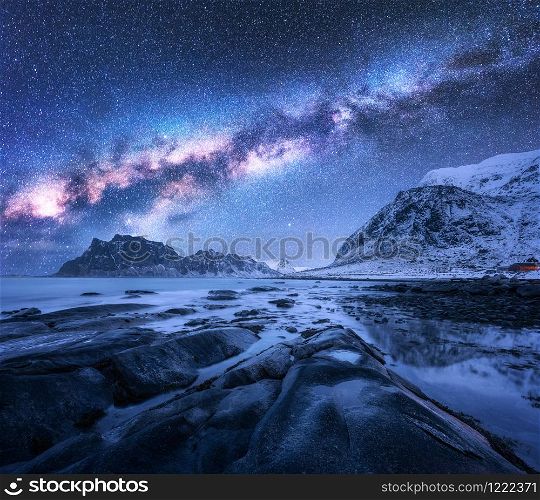 Milky Way over the snow covered mountains and rocky beach in winter at night in Lofoten Islands, Norway. Landscape with blue starry sky, water, stones, snowy rocks, bright galaxy. Beautiful space