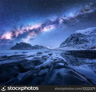 Milky Way over the snow covered mountains and rocky beach in winter at night in Lofoten Islands, Norway. Landscape with blue starry sky, water, stones, snowy rocks, bright galaxy. Beautiful space