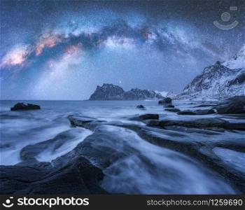 Milky Way over the snow covered mountains and rocky beach in winter at night in Lofoten Islands, Norway. Landscape with blue starry sky, water, stones, snowy rocks, bright milky way. Beautiful space