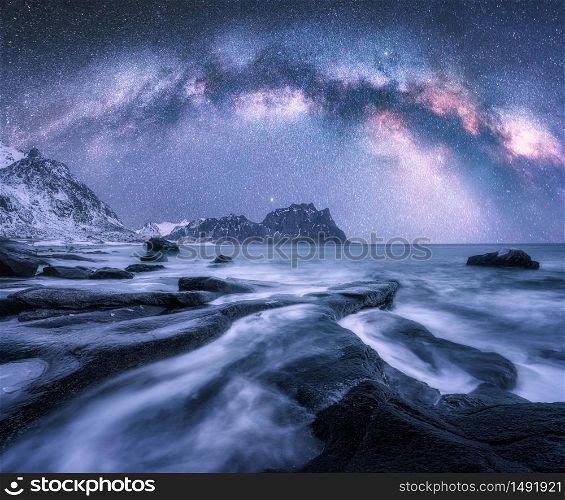 Milky Way over the snow covered mountains and rocky beach in winter at night in Lofoten Islands, Norway. Landscape with purple starry sky, water, stones, snowy rocks, bright milky way. Beautiful space