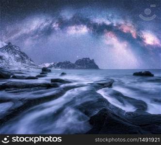 Milky Way over the snow covered mountains and rocky beach in winter at night in Lofoten Islands, Norway. Landscape with purple starry sky, water, stones, snowy rocks, bright milky way. Beautiful space