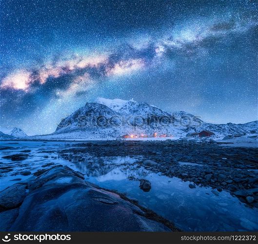 Milky Way over the snow covered mountains and rocky beach in winter at night. Lofoten Islands, Norway. Landscape with blue starry sky, water, stones, snowy rocks, bright galaxy and city lights. Space