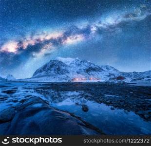 Milky Way over the snow covered mountains and rocky beach in winter at night. Lofoten Islands, Norway. Landscape with blue starry sky, water, stones, snowy rocks, bright galaxy and city lights. Space