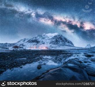 Milky Way over the snow covered mountains and rocky beach in winter at night. Lofoten Islands, Norway. Landscape with blue starry sky, water, stones, snowy rocks, bright galaxy and illumination. Space