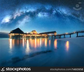 Milky Way over the wooden bungalow on the water at summer starry night. Landscape with hotel on the sea, lights, sandy beach, sky with stars and galaxy, reflection in water in Zanzibar, Africa. Space