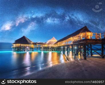 Milky Way over wooden bungalow on the water in summer starry night. Landscape with hotel on the sea, illumination, jetty, sandy beach, sky with stars, reflection in water in Zanzibar, Africa. Space