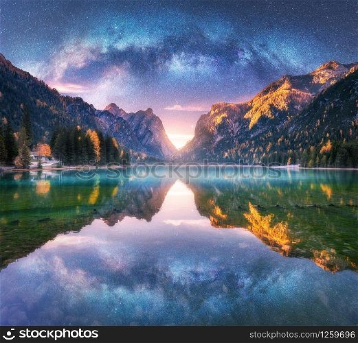 Milky Way reflected in water in mountain lake at starry night. Autumn landscape with purple sky with stars, blue fog, reflection, trees with colorful leaves, rocks in fall at sunset. Space and galaxy