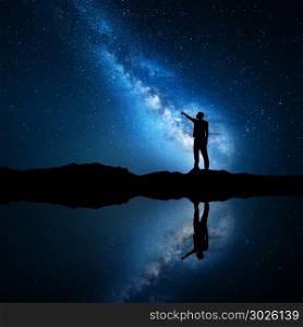 Milky Way. Silhouette of a standing man pointing finger in night starry sky on the mountain near the lake with sky reflection in water. Night landscape with universe. Blue milky way and man. Galaxy