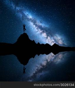 Milky Way. Silhouette of a standing woman practicing yoga on the mountain peak near the lake with sky reflection in water. Landscape with meditating girl on the rock. Night starry sky and milky way