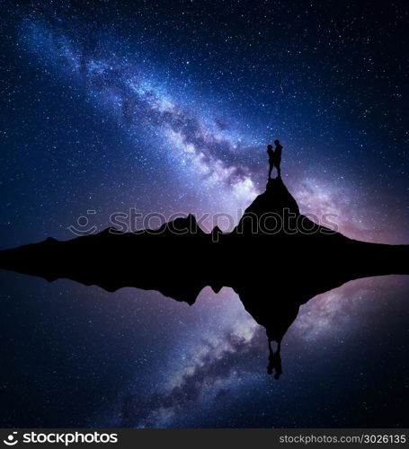 Milky Way. Silhouette of people. Landscape with night starry sky. Standing man and woman on the mountain peak near the lake with sky reflection in water. Hugging couple and milky way. Galaxy. Universe