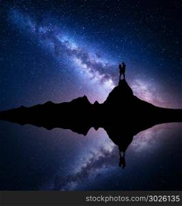 Milky Way. Silhouette of people. Landscape with night starry sky. Standing man and woman on the mountain peak near the lake with sky reflection in water. Hugging couple and milky way. Galaxy. Universe