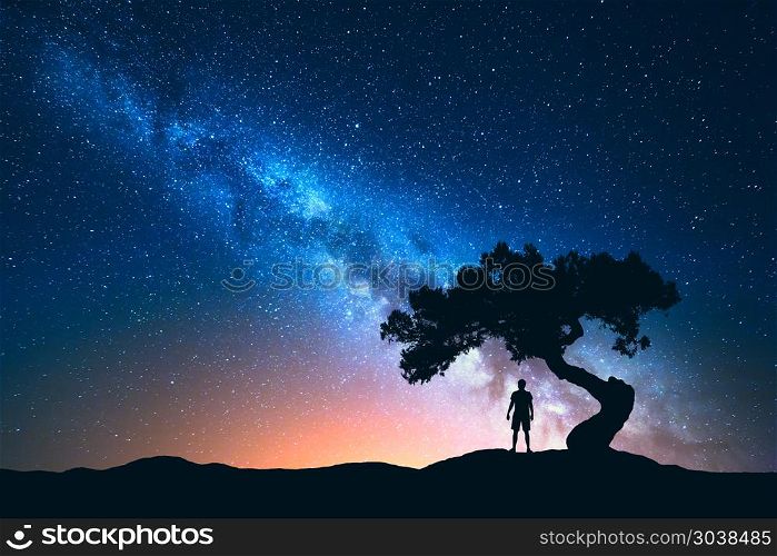 Milky Way, tree and silhouette of alone man. Night landscape. Night landscape with Milky Way and silhouette of man under the tree growing from the rock on the mountain. Nature background with starry sky and beautiful galaxy. Blue Milky Way and man. Universe