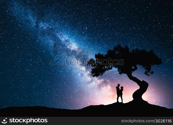 Milky Way with hugging couple under the tree on the hill. Landscape with night starry sky and silhouette of standing man and woman. Milky Way with young people. Space background. Amazing galaxy