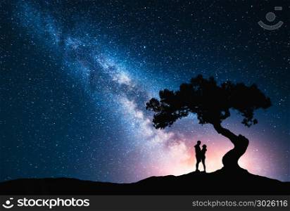 Milky Way with hugging couple under the tree on the hill. Landscape with night starry sky and silhouette of standing man and woman. Milky Way with young people. Space background. Amazing galaxy
