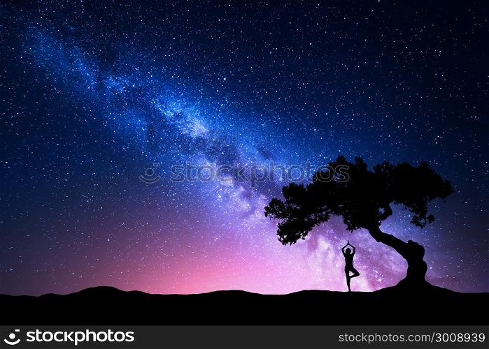 Milky Way with old tree and silhouette of a standing woman practicing yoga. Beautiful landscape with meditating girl under the tree against starry sky with pink milky way. Amazing galaxy. Universe