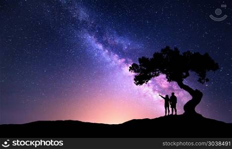 Milky Way with people under the tree on the hil. Milky Way with people under the tree on the hill. Landscape with night starry sky and silhouette of standing happy man and woman who pointing finger in starry sky. Milky way with travelers. Universe