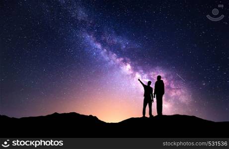 Milky Way with silhouette of a family and yellow city lights. Father and a son who pointing finger in night starry sky on the mountain. Night landscape. Beautiful Universe. Space. Travel background with sky full of stars