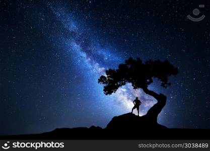 Milky Way with silhouette of a man. Milky Way. Night sky with stars, old tree and silhouette of a standing alone man with backpack on the mountain. Blue milky way and traveler. Travel background. Silhouette of a man under the tree