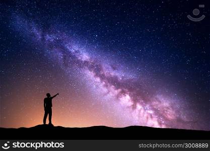 Milky Way with silhouette of a standing man pointing finger in night starry sky on the hill. Colorful night landscape. Beautiful Universe, travel background with purple sky yellow light. Space