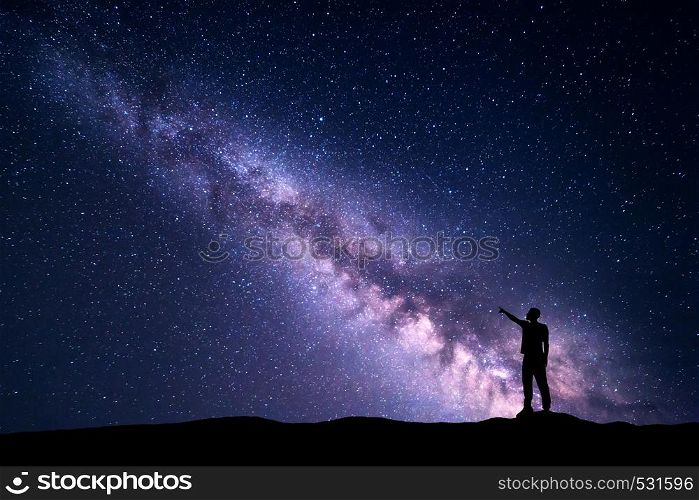 Milky Way with silhouette of a standing young man pointing finger in night starry sky on the mountain. Night landscape. Beautiful Universe, travel background with purple sky full of stars