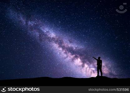 Milky Way with silhouette of a standing young man pointing finger in night starry sky on the mountain. Night landscape. Beautiful Universe, travel background with purple sky full of stars