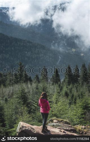 Millenial girl on a rock admiring the Hornisgrinde mountains covered by fir forest and clouds, in the Black Forest, Germany. Travel destination.