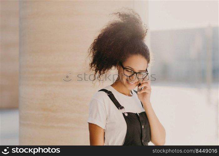 Millennial Afro woman speaks via cell phone, uses roaming connection, looks down, enjoys pleasant conversation, wears white t shirt and overalls, rests outdoors, calls via app, has good mood