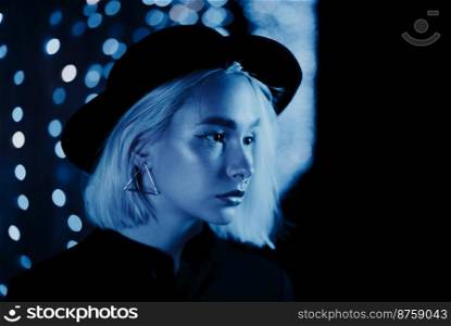 Millennial enigmatic pretty girl blond hairstyle near glowing neon wall at night. Blue hair, hipster hat, nose piercing. Mysterious woman. High quality photo. Millennial enigmatic pretty girl blond hairstyle near blue glowing neon wall at night. Hair, hipster hat, nose piercing. Mysterious woman