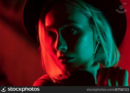 Millennial enigmatic pretty woman with blond dyed hairstyle near glowing neon wall at night. Nose piercing, hipster hat. Mysterious teenager. High quality photo. Millennial enigmatic pretty woman with blond dyed hairstyle near glowing neon wall at night. Nose piercing, hipster hat. Mysterious teenager.