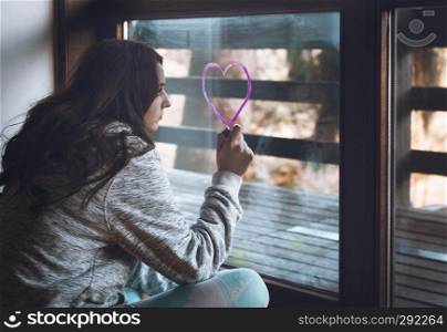 Millennial girl draws a heart with pink lipstick on a window.
