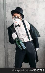 Mime actor with big bottle of alcohol performing a drunk man. Comedy pantomime artist in suit, gloves, glasses, make-up mask and hat. Mime actor performing a drunk man
