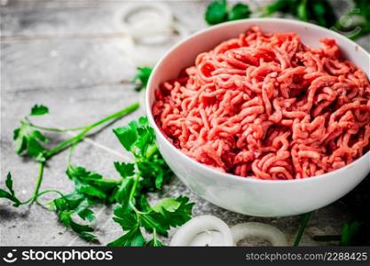 Minced meat in a bowl on a table with parsley and onion rings. On a gray background. High quality photo. Minced meat in a bowl on a table with parsley and onion rings.