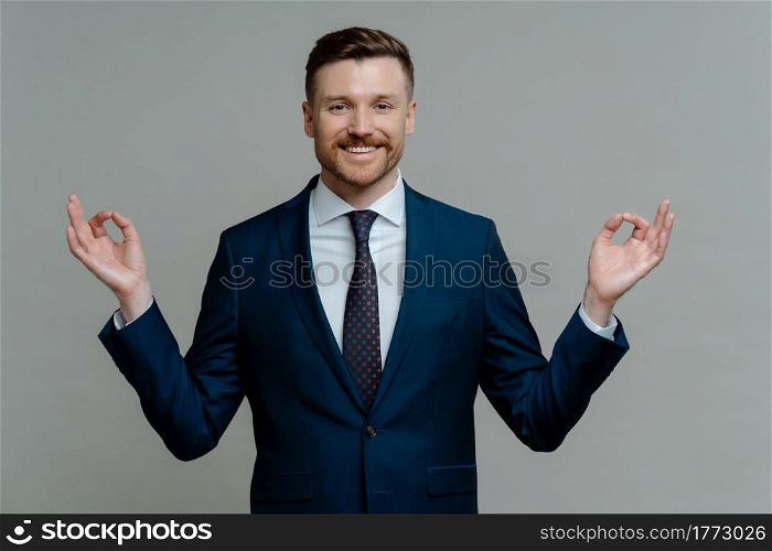 Mindful business people. Happy ceo executive in suit doing yoga exercise, successful businessman keeping hands in mudra and meditating, smiling at camera while standing against grey background. Happy businessman in suit meditating and feeling peaceful
