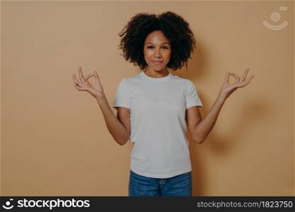 Mindfulness concept. Peaceful afro american young woman in casual outfit keeping hands in mudra gesture and looking at camera with tender smile, being calm and relaxed, isolated on beige wall. Peaceful afro american young woman keeping hands in mudra gesture, isolated on beige wall
