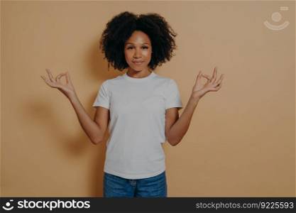 Mindfulness concept. Peaceful afro american young woman in casual outfit keeping hands in mudra gesture and looking at camera with tender smile, being calm and relaxed, isolated on beige wall. Peaceful afro american young woman keeping hands in mudra gesture, isolated on beige wall