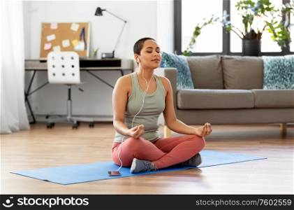 mindfulness, spirituality and healthy lifestyle concept - african american woman in earphones listening to music on smartphone and meditating in lotus pose at yoga studio. woman listening to music and meditating at tome