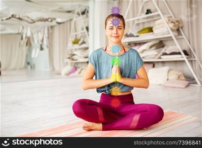 mindfulness, spirituality and healthy lifestyle concept - woman meditating in lotus pose at yoga studio with seven chakra symbols. woman meditating in lotus pose at yoga studio