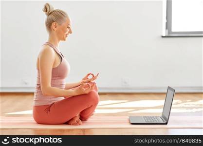 mindfulness, spirituality and healthy lifestyle concept - young woman with laptop computer meditating in lotus pose at yoga studio. woman with laptop in lotus pose at yoga studio