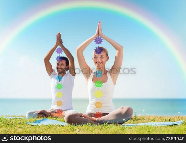 mindfulness, spirituality and outdoor yoga - couple meditating in lotus pose with seven chakra symbols over sea and rainbow background. couple doing yoga in lotus pose with seven chakras