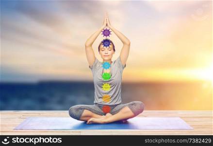 mindfulness, spirituality and outdoor yoga - woman meditating in lotus pose with seven chakra symbols over sea and sunlight background. woman doing yoga in lotus pose with seven chakras