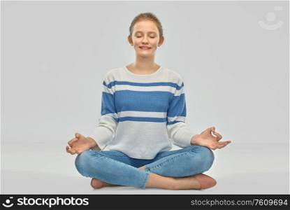 mindfulness, spirituality and people concept - smiling teenage girl in pullover meditating in lotus pose sitting on floor over grey background. smiling teenage girl meditating in lotus pose